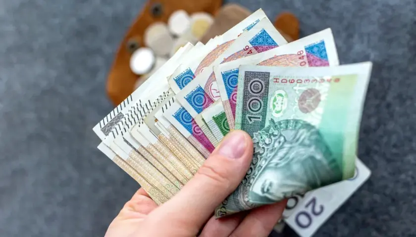 Dłoń trzymająca wachlarz polskich banknotów, w tle portfel z monetami.