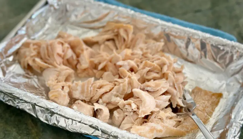 Flaked cooked salmon in a foil-lined baking dish, ready for a salmon flakes recipe. A fork rests on the side.