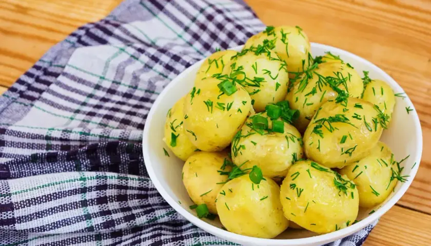 Młode ziemniaki w białej misce, posypane świeżym koperkiem i szczypiorkiem, na tle kratkowanej serwetki i drewnianego stołu.