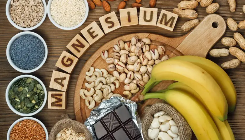 Magnesium-reiche Lebensmittel wie Sesam, Nüsse, Bananen und dunkle Schokolade sind auf einem Holztisch arrangiert.