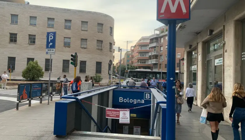 Ingresso della metro Bologna a Roma, con insegna rossa e bianca. Persone camminano sul marciapiede.