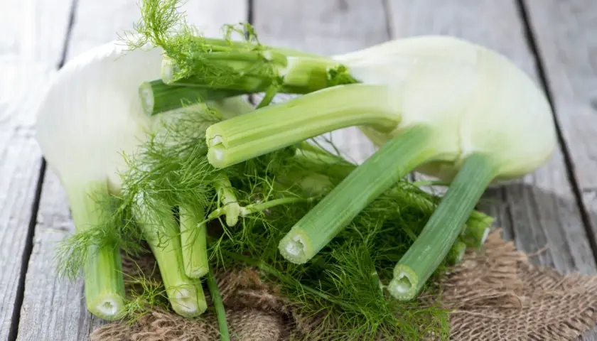 Kilka świeżych bulw fenkułu z zielonymi łodygami i liśćmi, ułożonych na drewnianym blacie. Fenkuł co to? To aromatyczne warzywo.