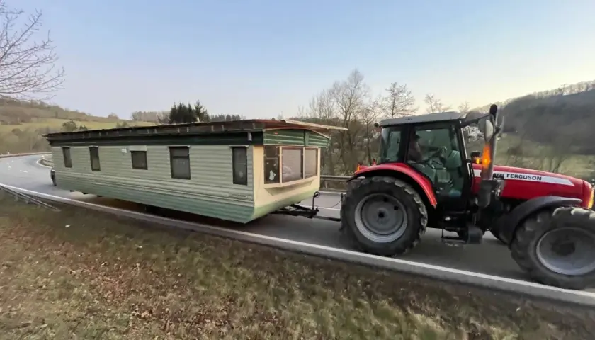 Ein roter Traktor zieht einen Wohnwagen über eine kurvige Straße. Sie sind mit Traktor und Wohnwagen unterwegs, um ein neues Zuhause zu finden.