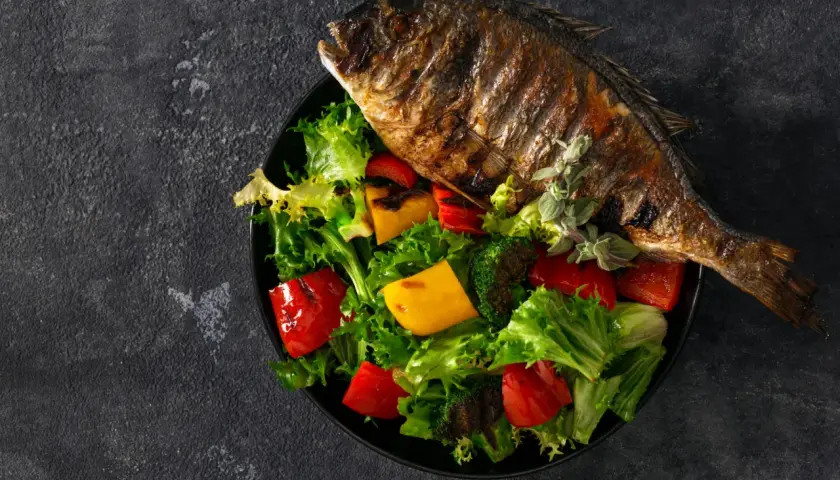 Dorade au barbecue, entière et grillée, servie sur un lit de salade verte, poivrons rouges et jaunes, et brocolis.
