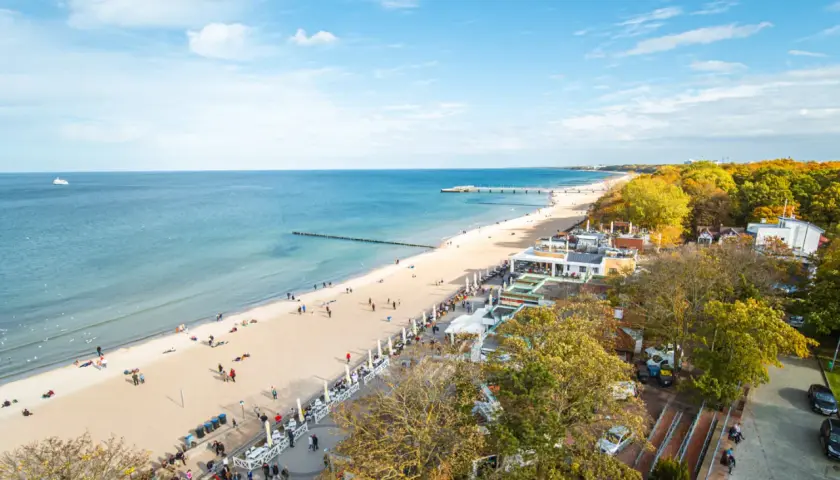 Słoneczny deptak w Kołobrzegu tętni życiem. Ludzie spacerują po plaży i promenadzie, ciesząc się jesiennym słońcem i widokiem na morze.