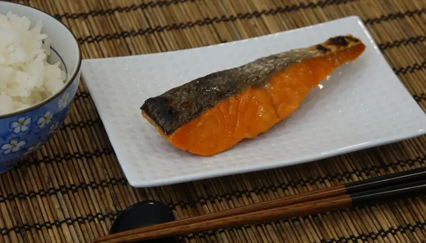 A perfectly grilled salmon shioyaki rests on a white plate, next to a bowl of white rice and chopsticks.