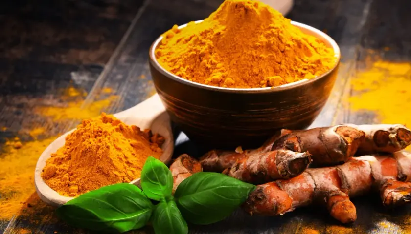 Radici di curcuma e polvere dorata in un cucchiaio e ciotola, con foglie verdi. La curcuma è nota per le sue proprietà antinfiammatorio.