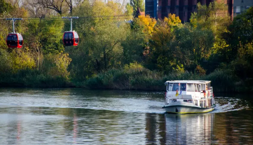 Dwie czerwone gondole kolejki linowej "Polinka" nad Odrą we Wrocławiu, obok płynie statek.