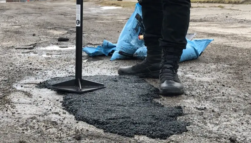 Pracownik ubija asfalt na zimno, naprawiając dziurę w drodze. Obok leży niebieski worek z materiałem.