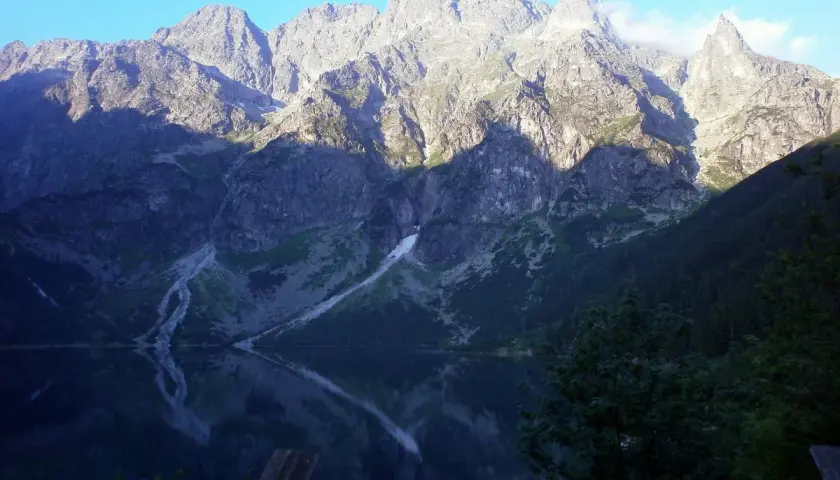 Morskie Oko odbija się w spokojnej tafli wody. Majestatyczne góry, oświetlone słońcem, wznoszą się na tle błękitnego nieba. Wysokość nad poziomem morza robi wrażenie.