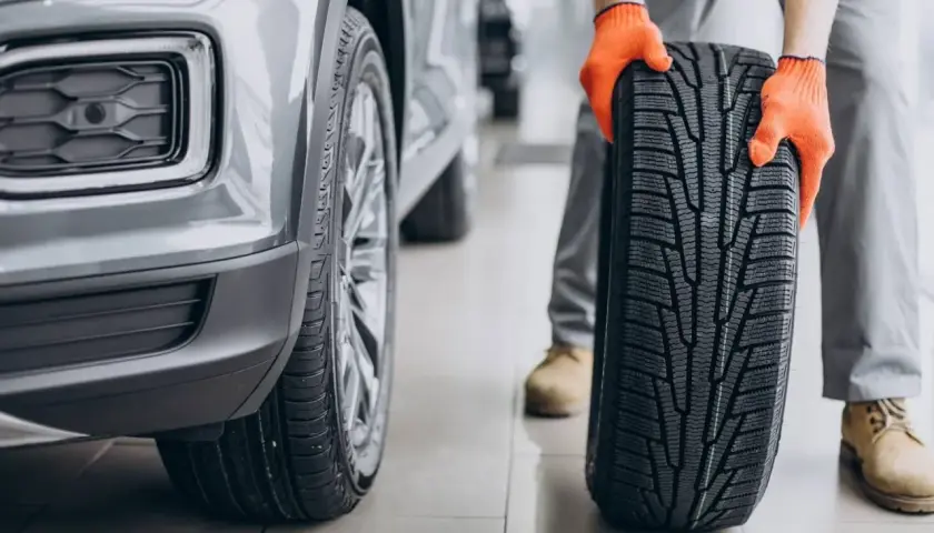 Mechanik w pomarańczowych rękawicach prezentuje nową oponę asymetryczną obok samochodu.