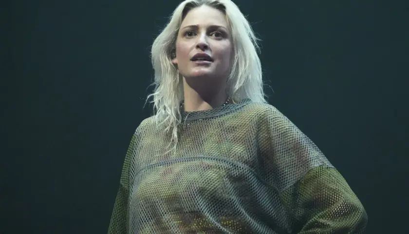 Blond wokalistka w siatkowej bluzce, przypominająca styl wokalisty Linkin Park, na ciemnym tle.