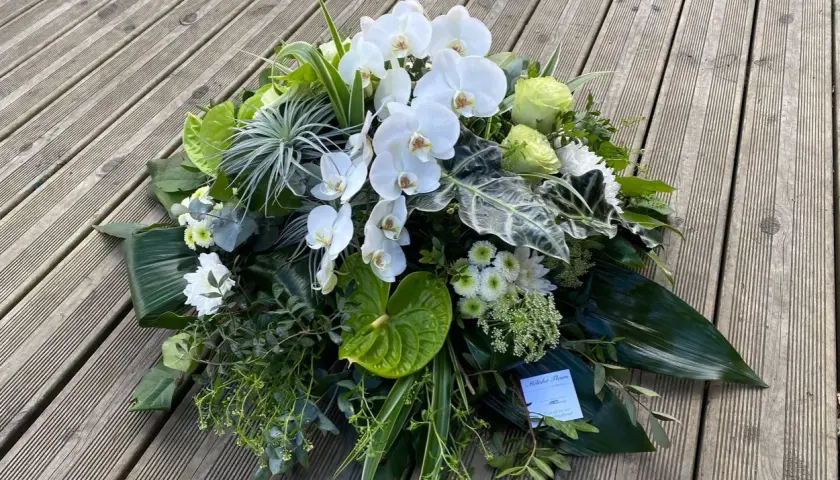 Un arrangement floral blanc et vert, composé d'orchidées, de roses et d'anthuriums, évoque un coussin de deuil moderne.