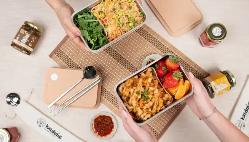 Two hands hold separate noodle bento boxes, one with fried rice and greens, the other with noodles, strawberries, and orange slices.