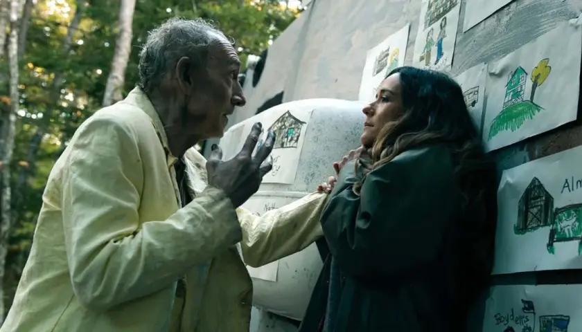 Un vieil homme aux cheveux gris, vêtu d'une chemise jaune, parle à une femme. Des dessins d'enfants ornent le mur derrière eux. Saison 5 date de sortie.