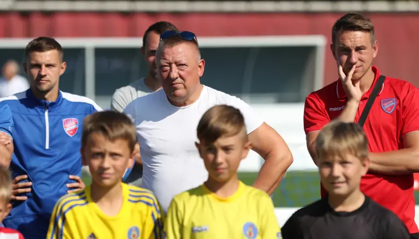 Marek Śledź, trener, w białej koszulce, stoi wśród młodych piłkarzy na boisku.