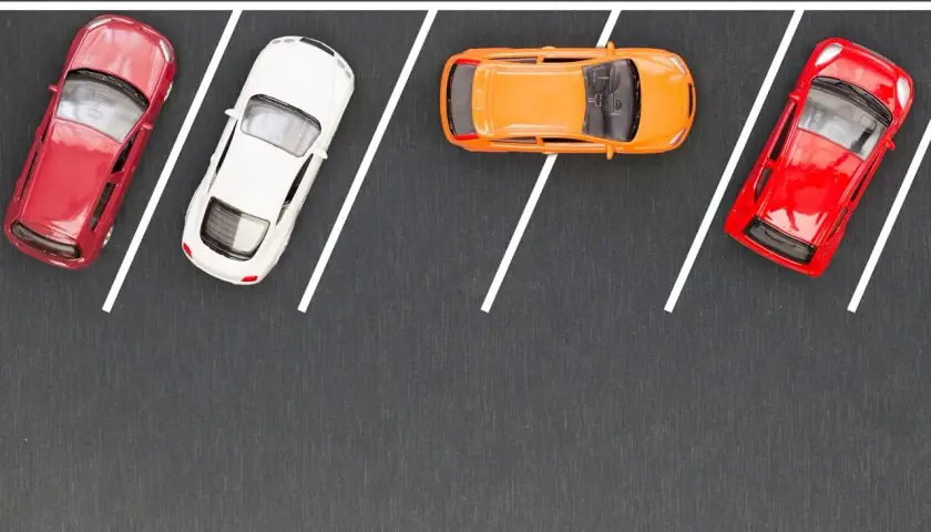 Cztery samochodziki: czerwony, biały, pomarańczowy i ponownie czerwony, zaparkowane na szarym tle z białymi liniami wyznaczającymi miejsca parkingowe.