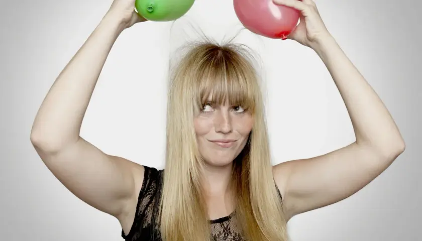En kvinna med blont hår och fräknar håller upp en grön och en röd ballong ovanför huvudet, vilket får hennes hår att resa sig av statiskt elektricitet.