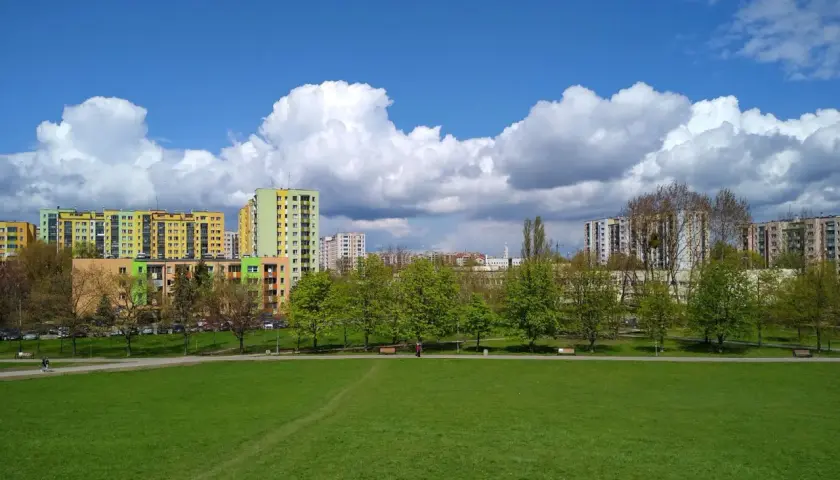Zielona przestrzeń w parku Górczewska, z kolorowymi blokami mieszkalnymi w tle i białymi chmurami na błękitnym niebie.