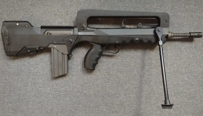 Czarny karabin FAMAS z charakterystycznym uchwytem i dwójnogiem, leżący na szarej tkaninie.