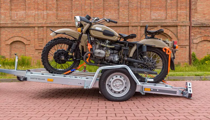 Motocykl Ural na przyczepie z pasami transportowymi, na tle ceglanego budynku.