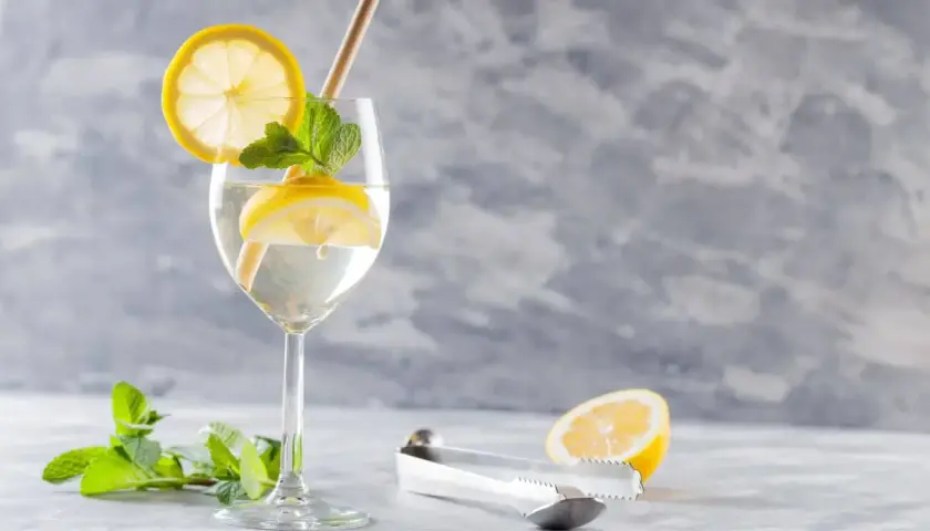 Szklanka z drinkiem Hugo, plasterkami cytryny, miętą i słomką. Obok leży połówka cytryny i szczypce.