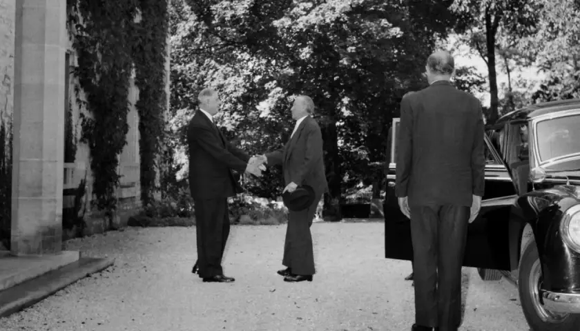 Konrad Adenauer und Charles de Gaulle schütteln sich die Hände. Ein Mann im Anzug steht daneben.
