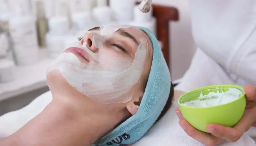 Une femme reçoit un masque ou gommage en premier, appliqué au pinceau sur son visage. Elle est détendue, les yeux fermés.
