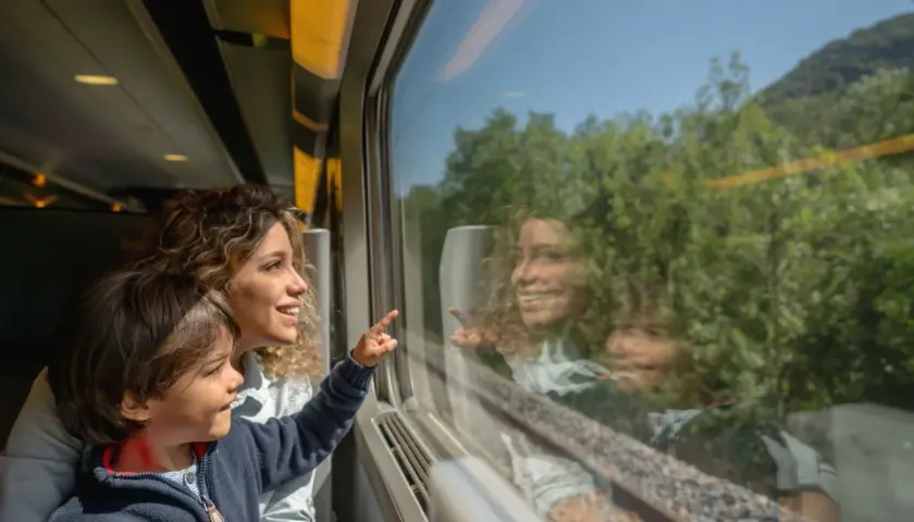 Bambino e madre guardano fuori dal finestrino del treno, indicando qualcosa. Un'ottima idea per i giochi da fare in treno, osservando il paesaggio che scorre.