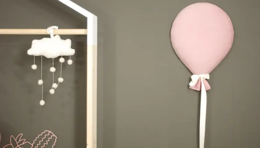Różowy balon z kokardką przyczepiony do szarej ściany obok dziecięcej dekoracji.