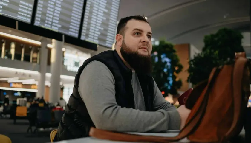 Mężczyzna z brodą siedzi w poczekalni lotniska, patrząc na tablicę odlotów. Czyżby czekał na lot, który jest zagrożony przez overbooking?