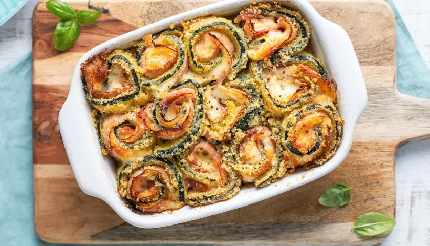 Involtini di zucchine al forno, dorati e filanti, con prosciutto e formaggio, guarniti con basilico fresco.