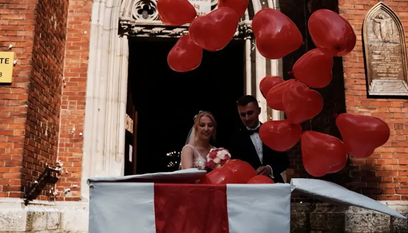 Para młoda wychodzi z kościoła, a rodzice przygotowali niespodziankę: mnóstwo czerwonych balonów w kształcie serc.