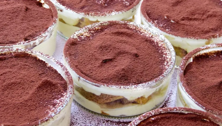 Varios vasitos de tiramisú, con capas de crema y bizcocho, espolvoreados con cacao. Los tiramisú ingredientes se ven deliciosos.
