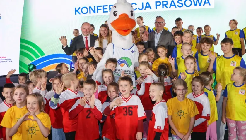 Uśmiechnięte dzieci i duża maskotka z logo lekkoatletycznych mistrzostw świata. Wszyscy pozują do zdjęcia, w tle napis "Konferencja prasowa".