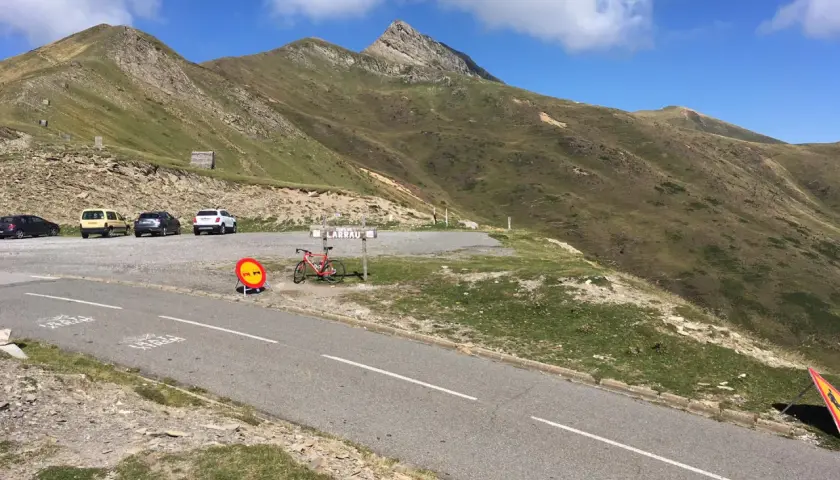Un ciclista descansa junto a su bicicleta roja en el **puerto de Larrau**, con coches aparcados y montañas verdes bajo un cielo azul.