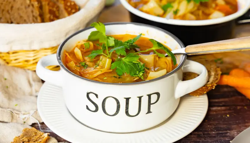 Herzhafte Kohlsuppe, low carb, in einem weißen Topf mit der Aufschrift "SOUP", garniert mit Petersilie. Dazu gibt es Vollkornbrot und Karotten.
