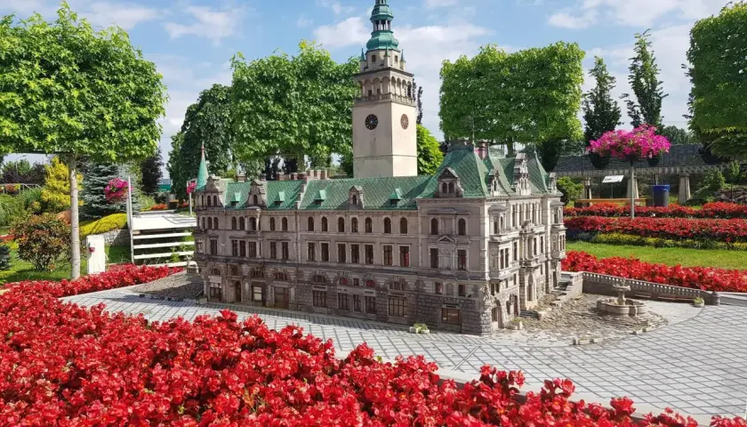 Miniatura ratusza z wieżą zegarową, otoczona czerwonymi kwiatami, w parku miniatur w Kotlinie Kłodzkiej.