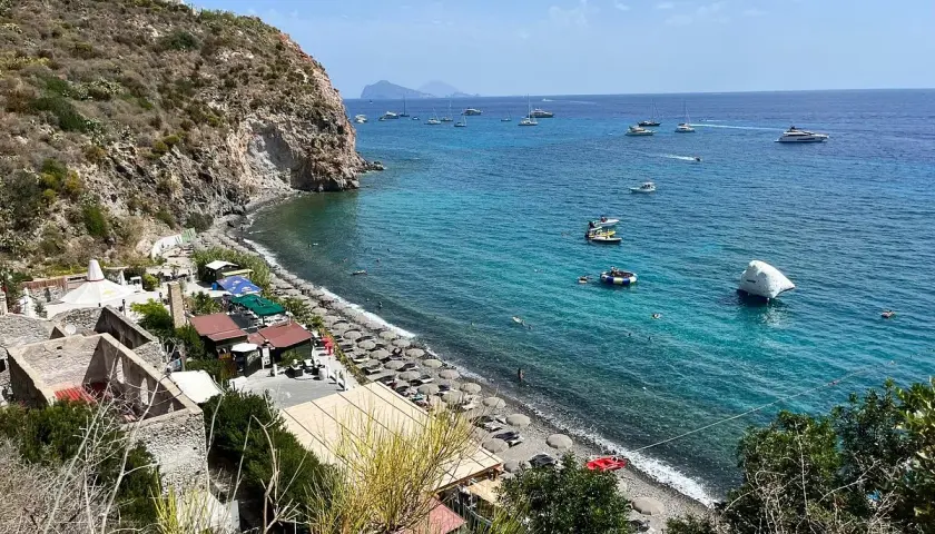 Plage animée à Lipari, avec des parasols, des petits restaurants et des bateaux naviguant sur la mer turquoise.