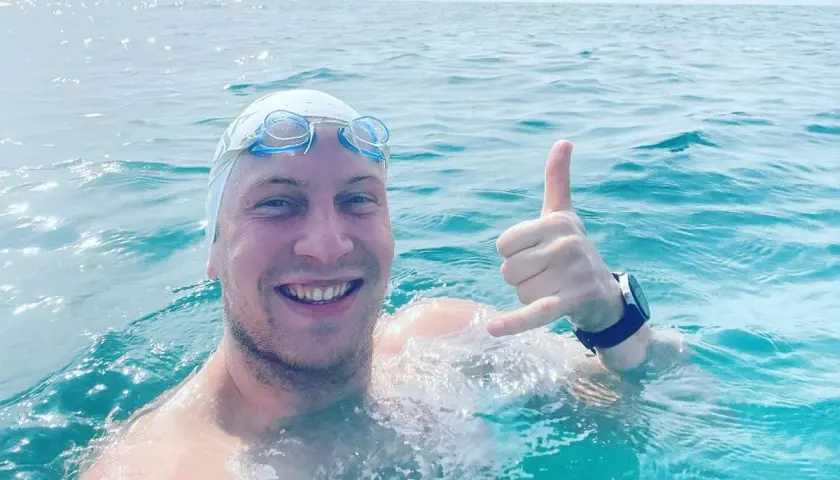 Uśmiechnięty pływak w czepku i goglach, z ręką w geście "hang loose", w trakcie przepłynięcia Bałtyku wpław.