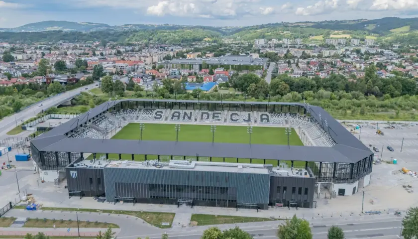 Nowoczesny stadion Sandecji z zieloną murawą i trybunami, otoczony miastem i wzgórzami.