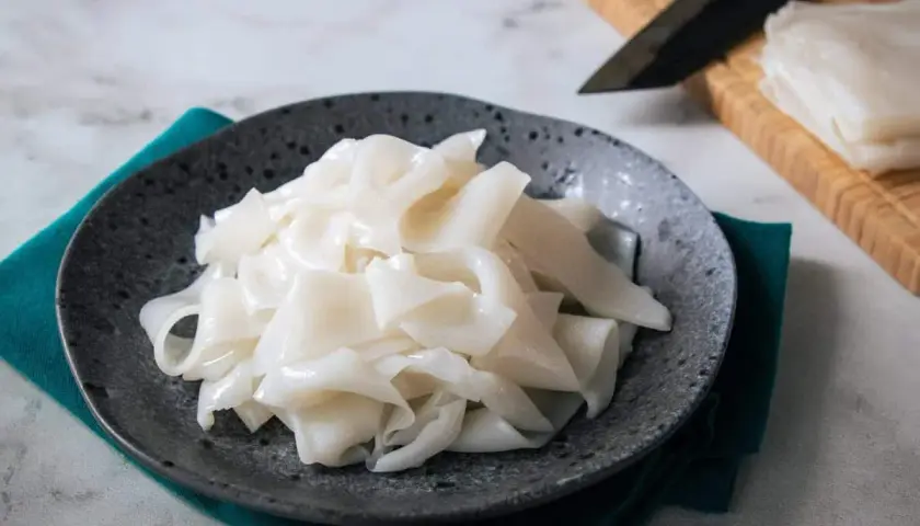 Na talerzu ułożone są białe, szerokie wstążki makaronu, przypominające capelli d'angelo. Obok leży nóż i deska do krojenia.