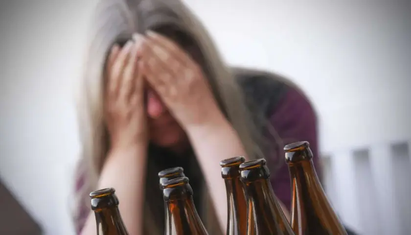 Verzweifelte Frau mit Händen vor dem Gesicht, umgeben von Bierflaschen. Ein Symbol für die Gefahren von Alkohol und die mögliche Entwicklung von Alkohol-Demenz.