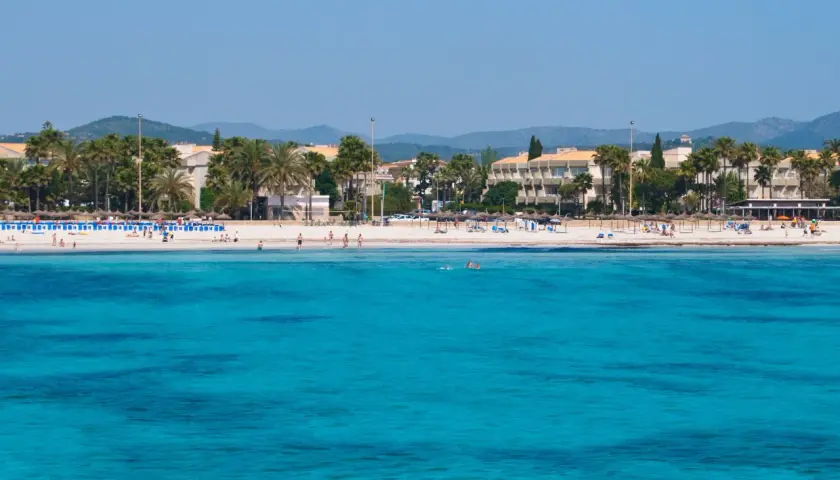 Türkisblaues Meer vor Sa Coma Altstadt mit Palmen und Hotels. Menschen genießen den Strand und das Wasser.