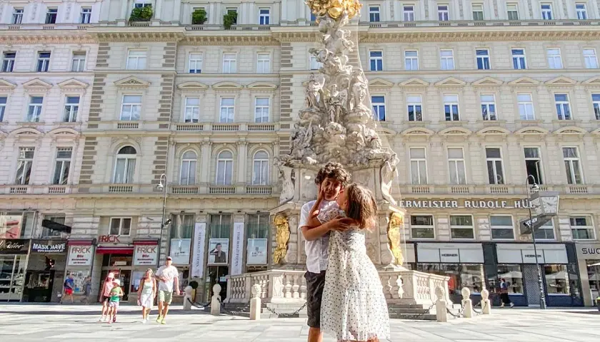 Due bambini si abbracciano davanti alla Colonna della Peste a Vienna. Altri bambini passeggiano sullo sfondo.