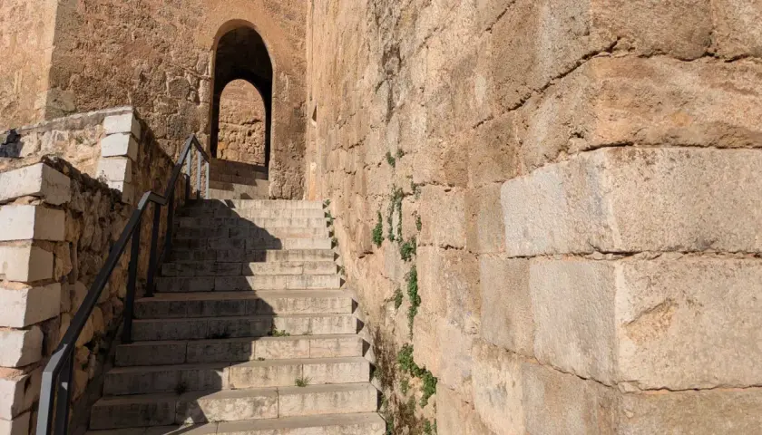 Escaleras de piedra ascienden hacia un arco en el castillo de Requena. La antigua muralla de piedra, con vegetación creciendo en sus grietas, proyecta sombras.