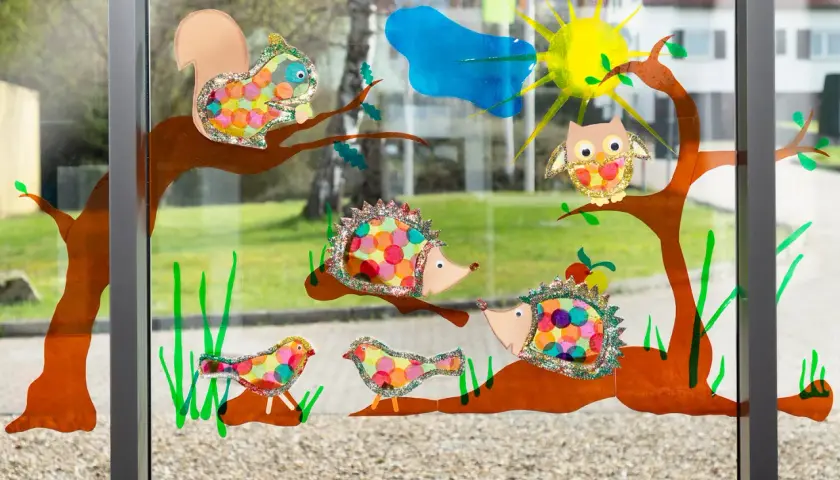 Bunte Fensterbilder basteln Kinder: Eichhörnchen, Igel, Eule & Vögel auf Ästen. Sonnenschein & Wolke im Hintergrund.