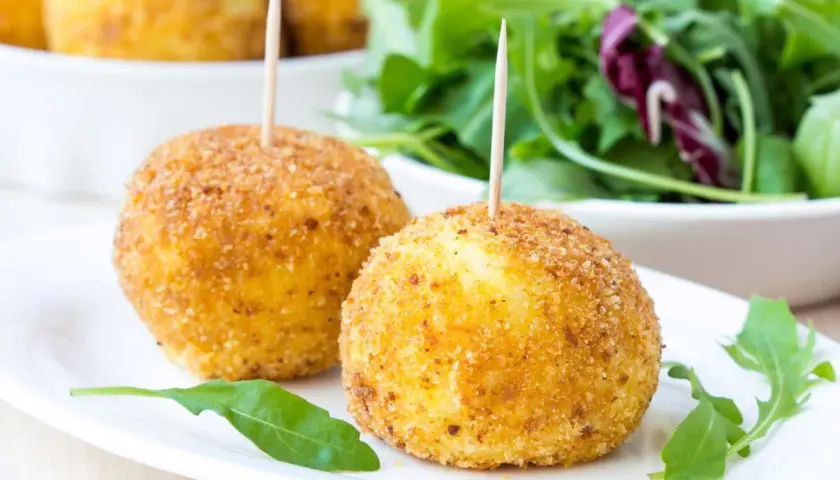 Dwa złociste arancini z wykałaczkami na białym talerzu, obok sałatka.