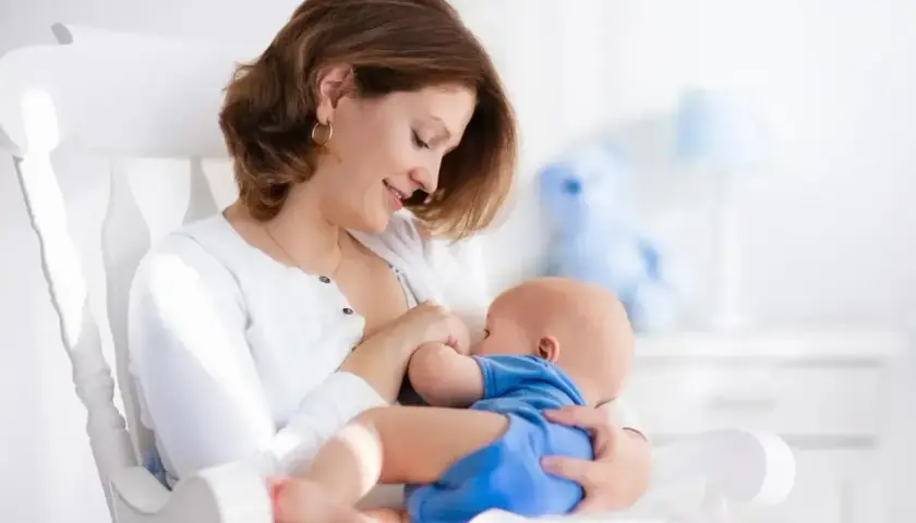 Uśmiechnięta mama karmi piersią noworodka w białym fotelu. Delikatne chwile bliskości, choć czasem pojawia się ulewanie u noworodka.