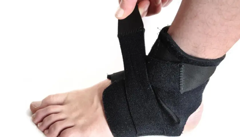 Dłoń zapina czarny stabilizator na kostce. Zapewnia wsparcie i ochronę podczas aktywności.